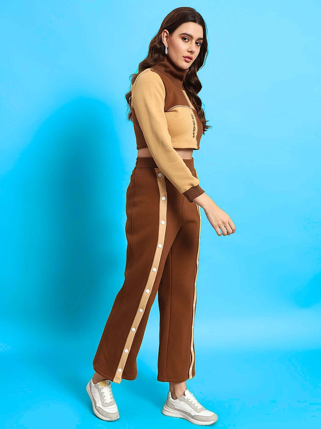 Woman wearing Women's Aurora Co-Ord Set in beige-brown, standing against blue background, showcasing stylish top and pants made from premium fabric.