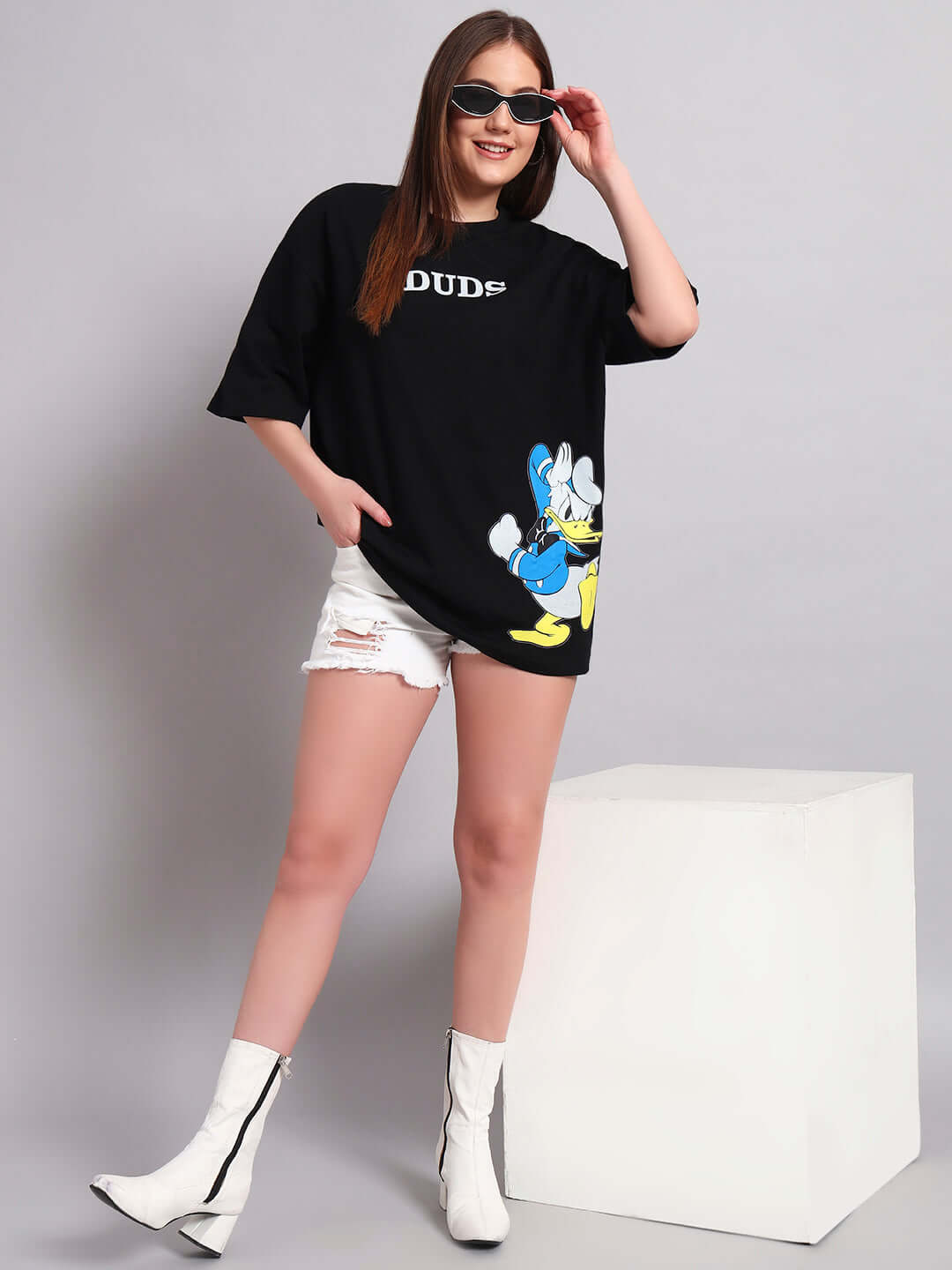 Woman wearing a black oversized T-shirt with Donald Duck print, styled with white shorts and boots, against a minimalist background.