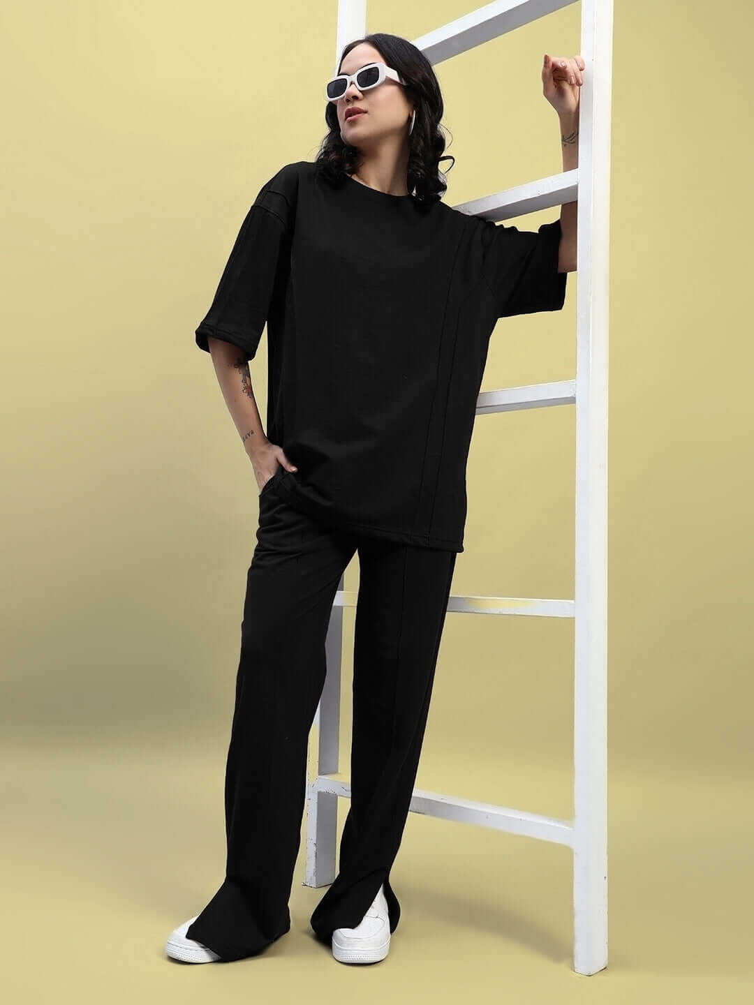 Woman wearing a black oversized T-shirt and pants, posed against a ladder, showcasing fashion style.