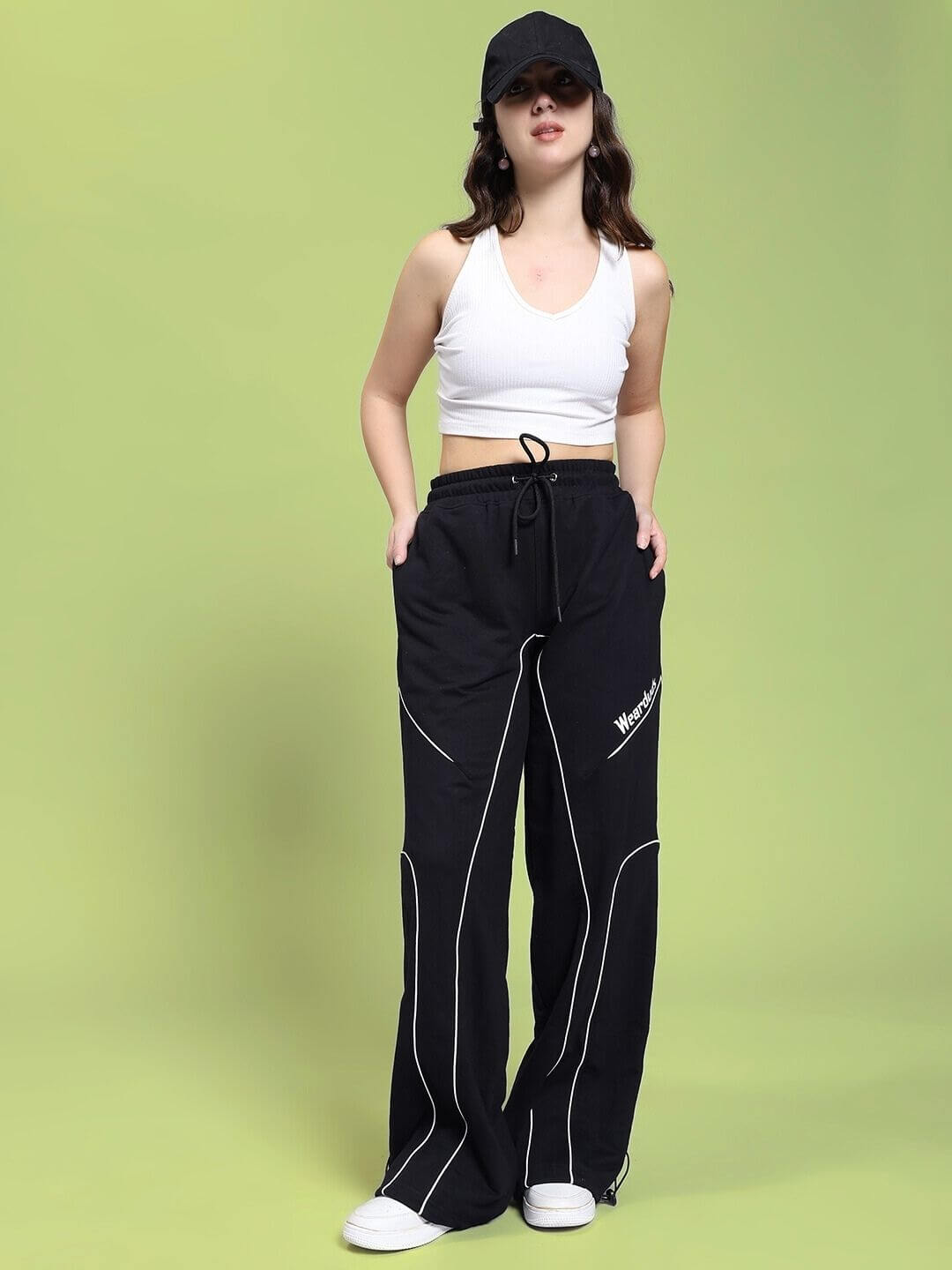 Woman wearing WOMEN SIGNATURE PIPING JOGGERS BLACK with contrast piping and a white crop top against a green background.