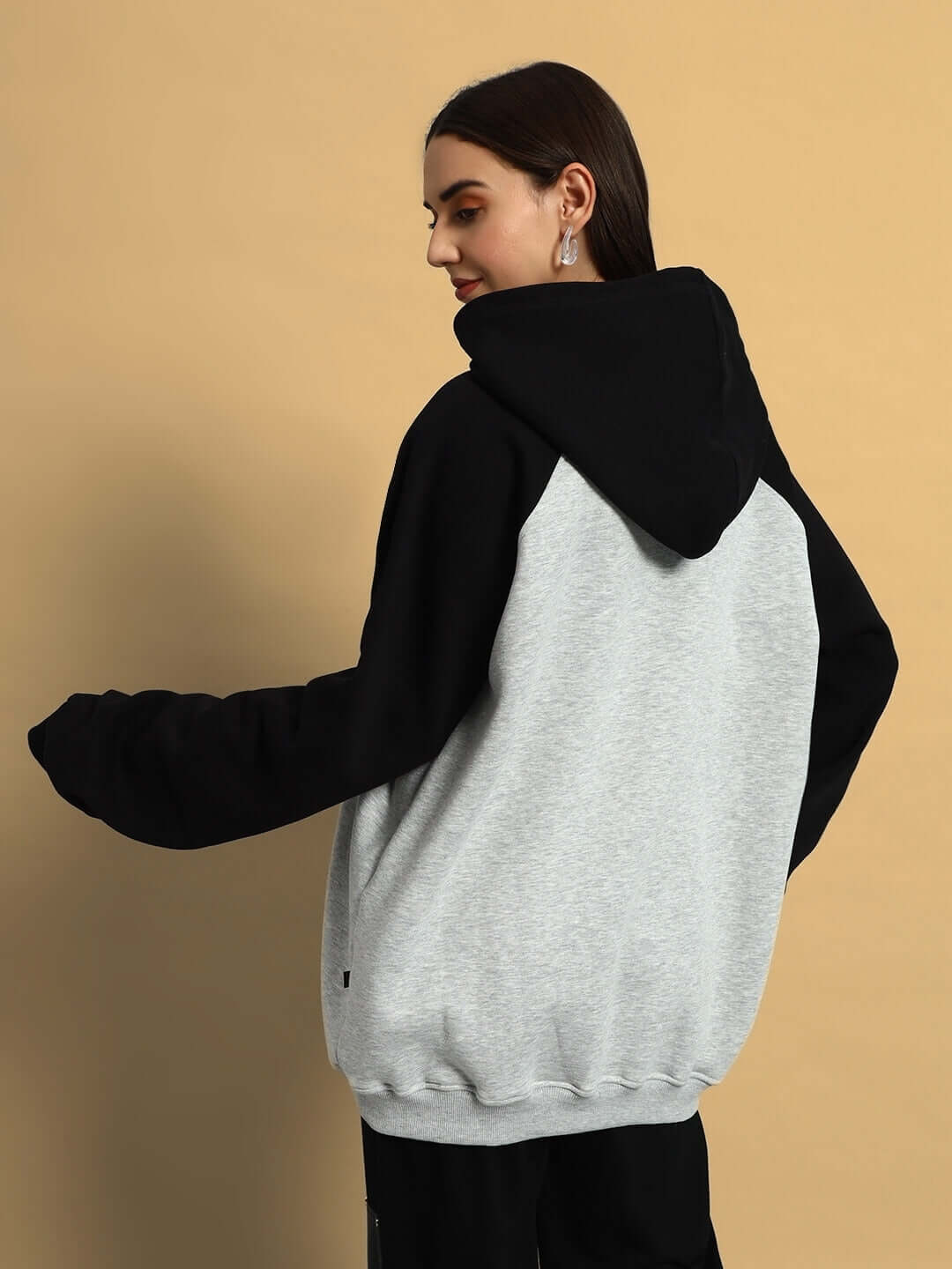 Woman wearing WOMEN DELUXE FLECCE OVERSIZE HOODIE BLACK MELANGE GREY, showcasing back view and relaxed fit against beige background.