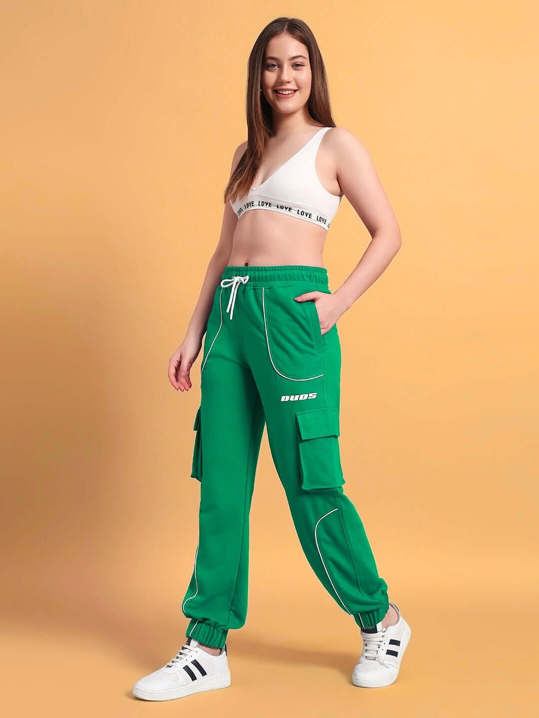 Woman wearing green relaxed-fit joggers with cargo pockets, styled with a white sports top, displaying casual chic fashion on a beige backdrop.
