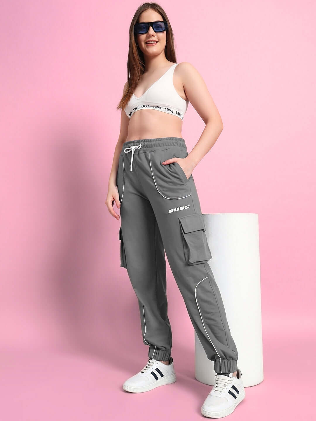 Woman wearing grey relaxed-fit joggers with pockets, styled with casual sneakers and a white crop top against a pink background.