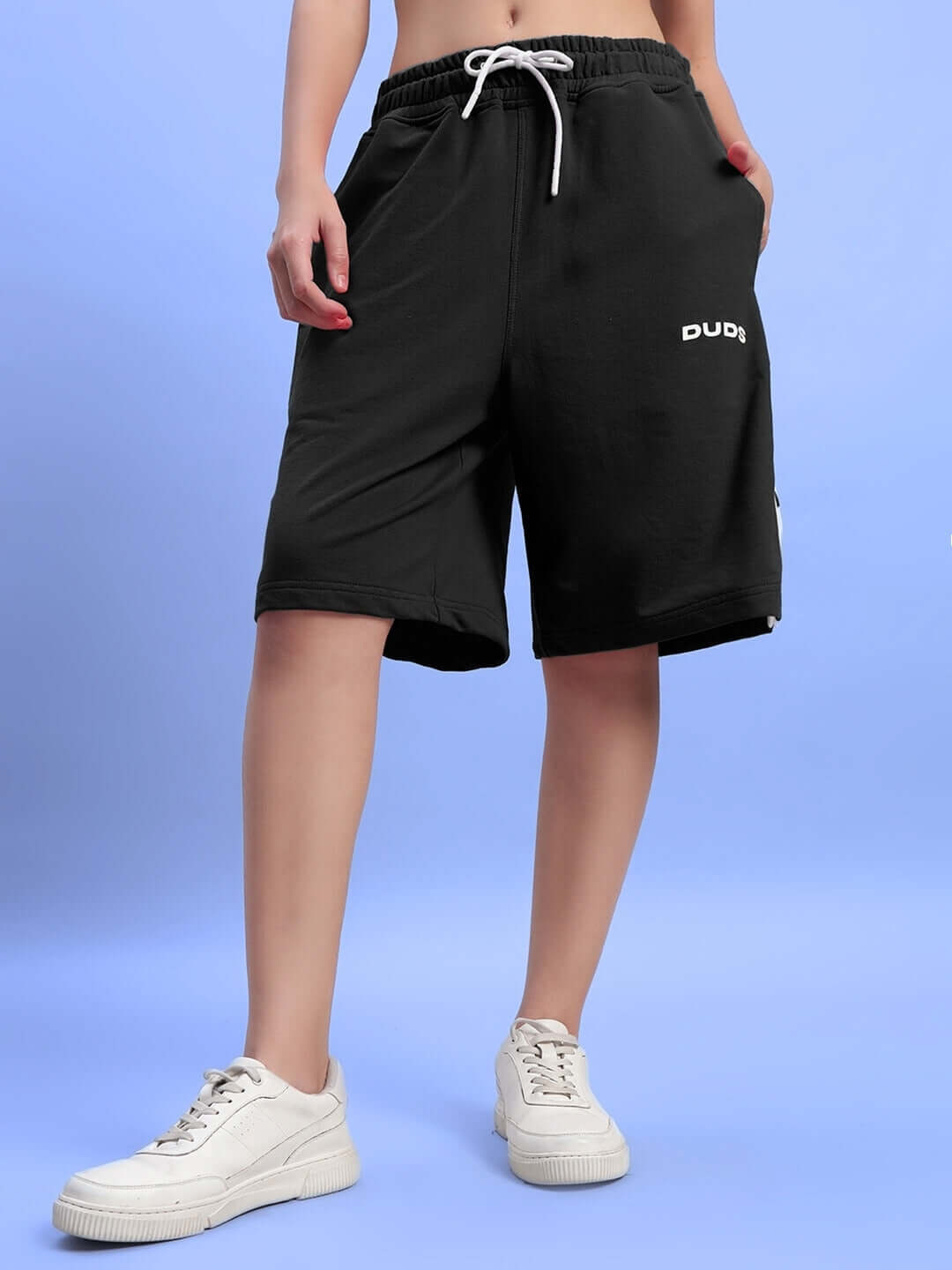 Woman wearing black Ezel regular fit shorts by DUDS with white drawstring, standing on blue background. Casual summer wear.