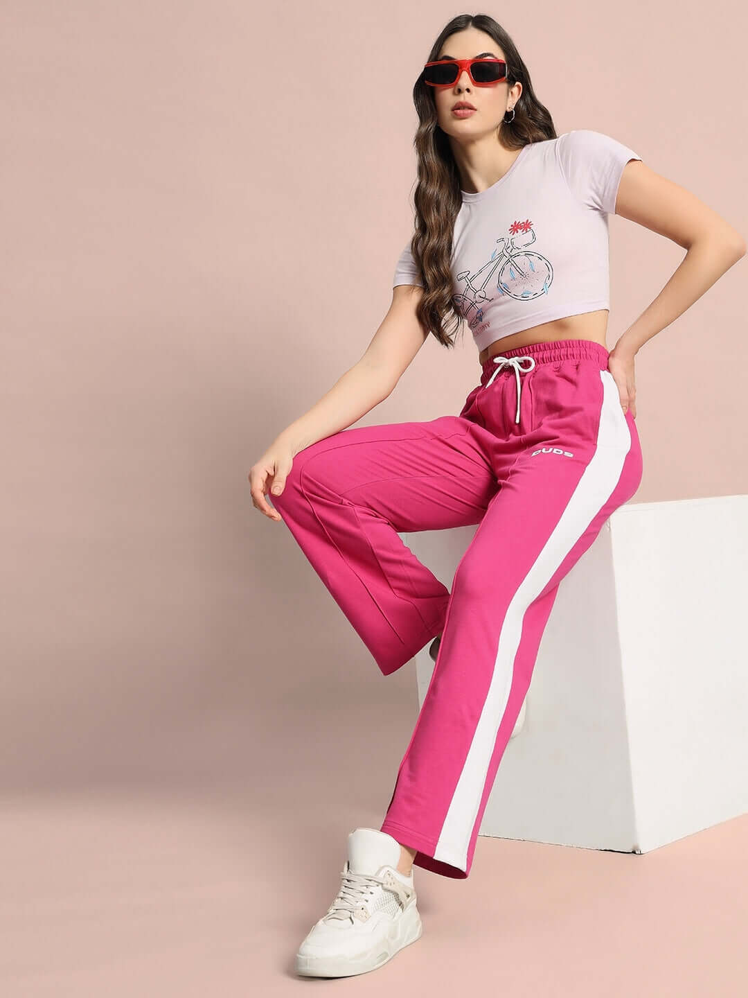 Model wearing lilac women's crop-top and pink track pants, styled with white sneakers and sunglasses, posing against a peach background.