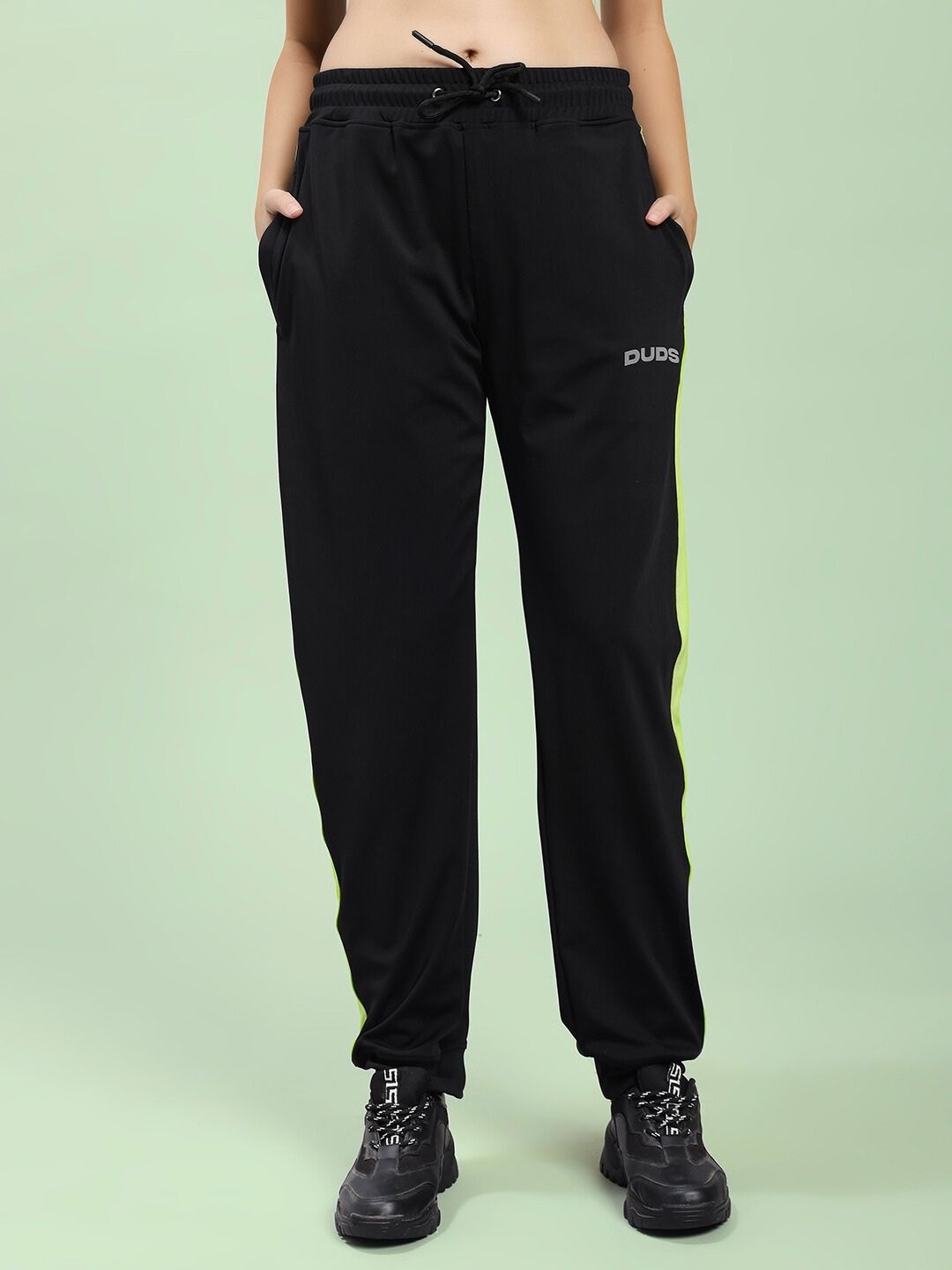 Model wearing WOMEN HIGH PERFORMANCE CO-ORD SET BLACK with tapered joggers and sneakers on a green background.