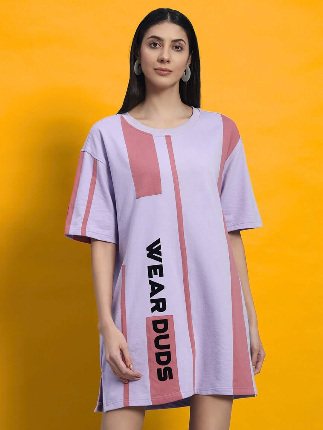 Model wearing lilac oversized t-shirt dress with "WEAR DUDS" design, standing against a yellow background.