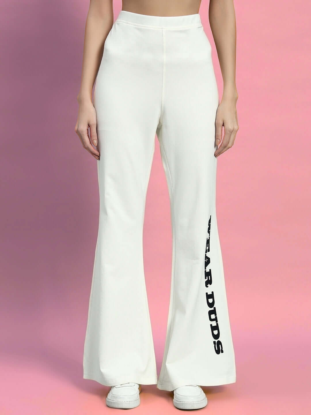 Woman wearing off-white relaxed fit joggers with elastic waistband, featuring "WEAR DUDS" text on the leg, against a pink background.