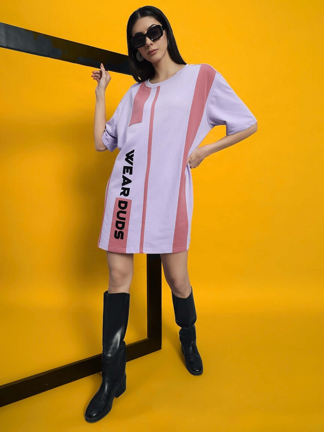 Model wearing stylish lilac oversized dress with "WEAR DUDS" lettering, black boots, and sunglasses against a yellow background.
