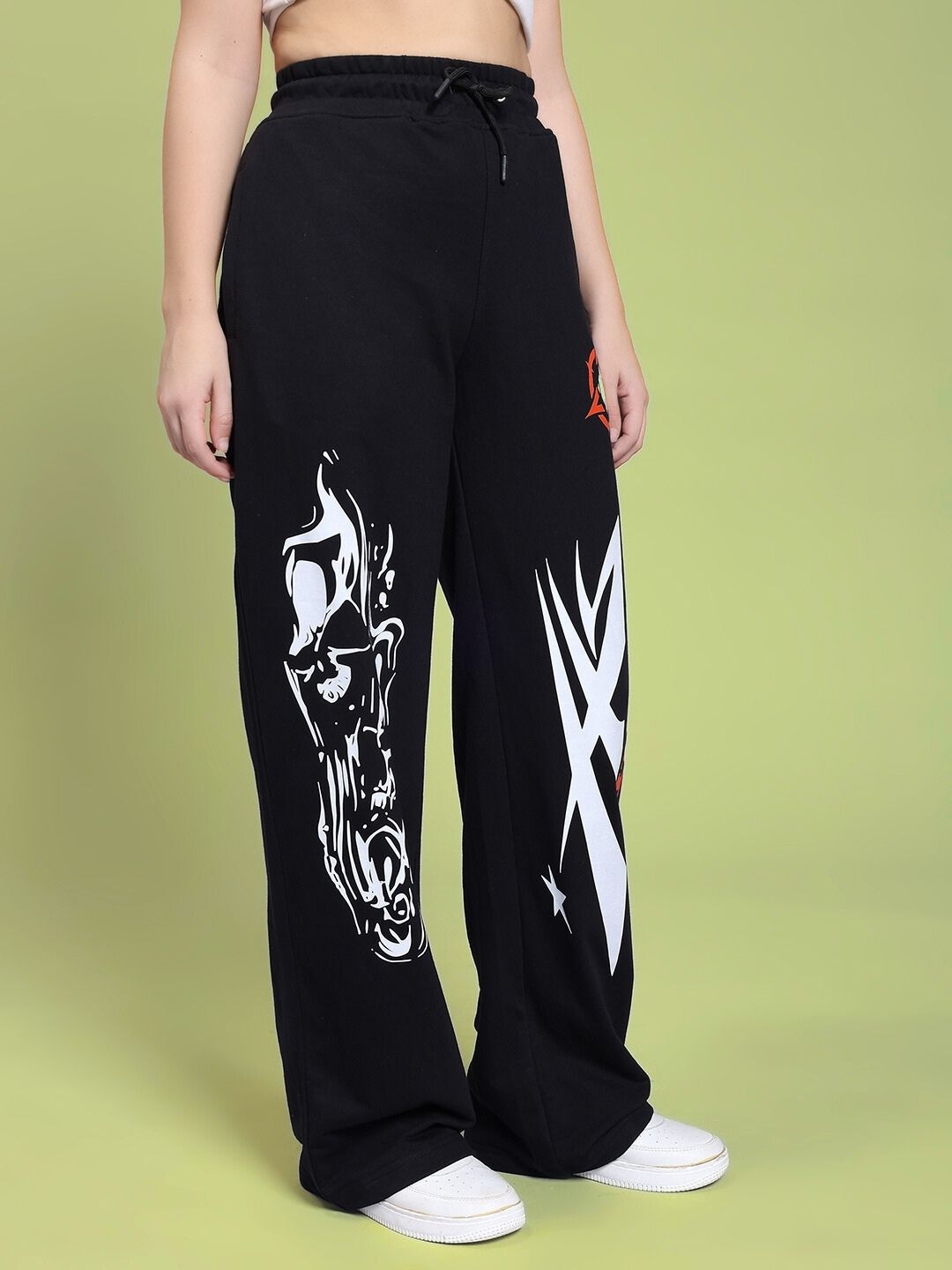 Model wearing WOMEN RIFT RELAXED FIT JOGGER IN BLACK with white sneakers against a green background, showcasing stylish prints.