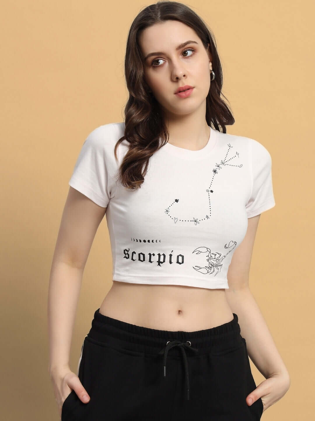 Woman wearing WOMEN SCORPIO CROP-TOP BABY PINK with Scorpio print, standing against beige background.