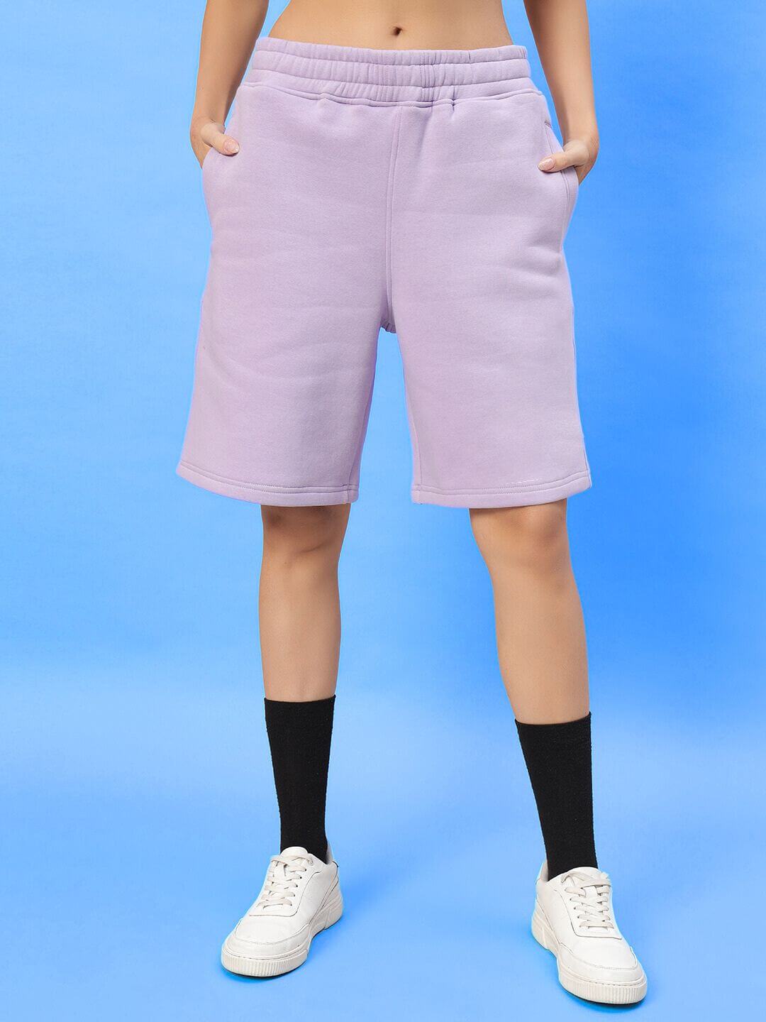 Woman wearing WOMENS SILENT HUG PRINTED FLEECE CO-ORD LILAC shorts with sneakers on blue background.