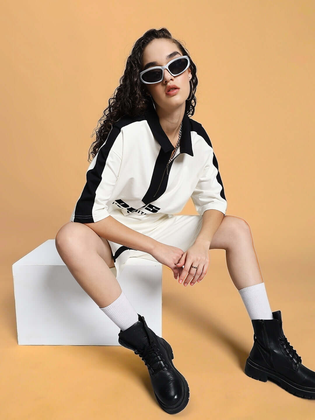 Model wearing WOMEN SPLINTER SUMMER CO-ORD SET OFF WHITE with black boots and sunglasses, seated against a beige background.