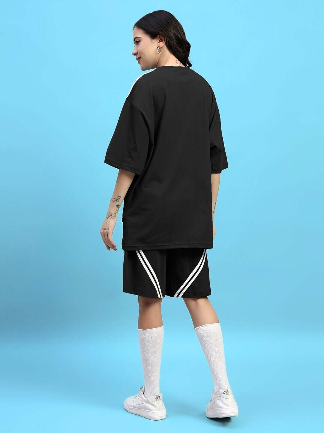 Back view of woman in black Women's Star Jumper Regular Fit Shorts with white stripes, paired with white socks and sneakers against blue background.