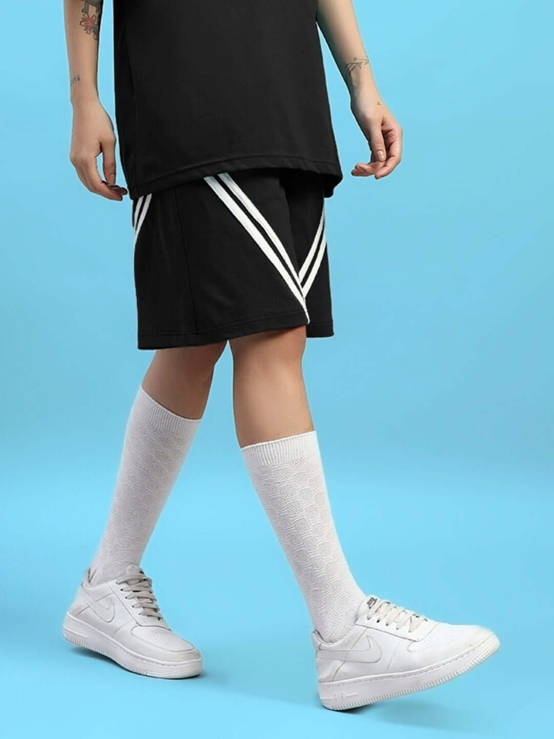 Model wearing WOMEN'S STAR JUMPER regular fit black shorts with white sneakers, standing against a blue background.