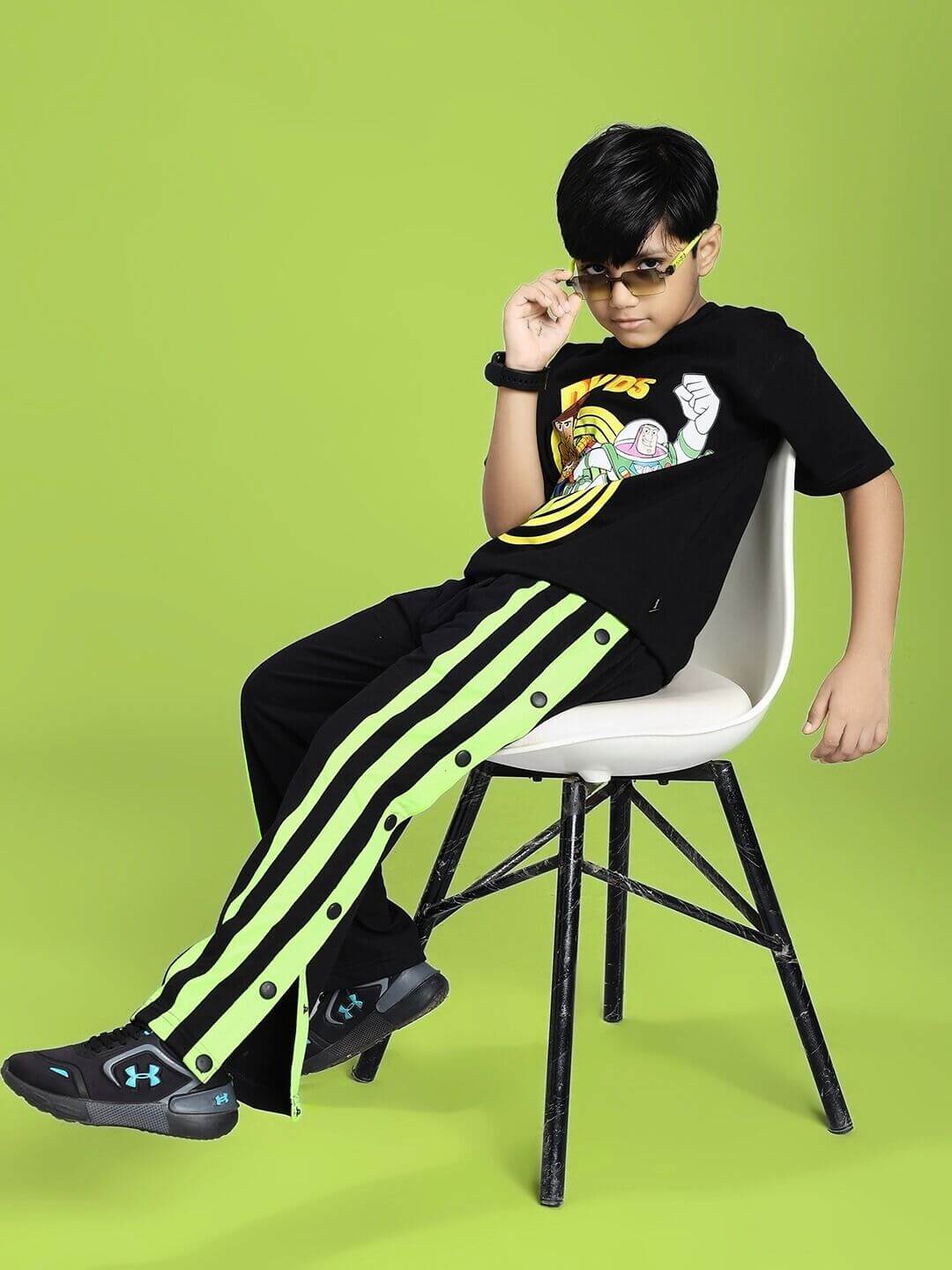 Child wearing WOODY BUZZ T-SHIRT FOR BOYS AND GIRLS BLACK with character graphic, sitting stylishly on a chair, green background.