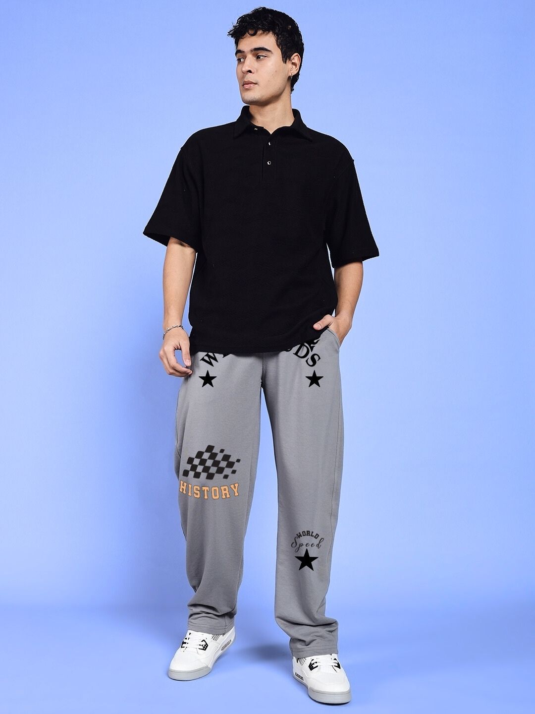 Man wearing WORLD SPEED PRINTED RELAXED FIT JOGGERS GREY with black shirt and sneakers, standing against a blue background.