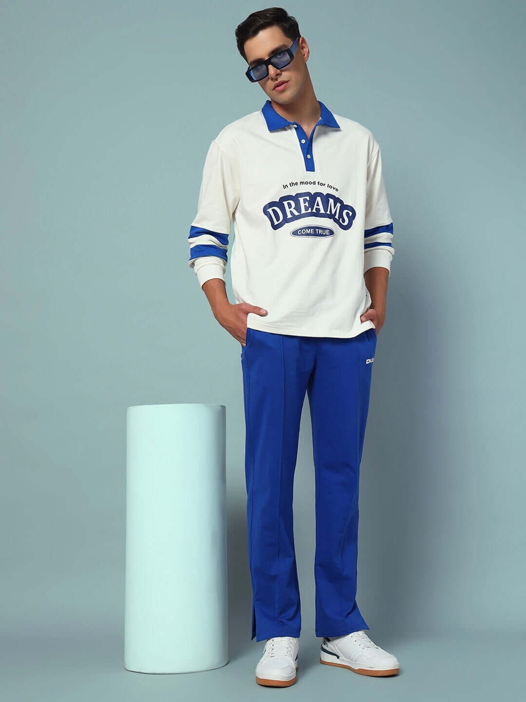Model wearing ZION SIDE SLIT JOGGERS in royal blue, styled with a casual white and blue long-sleeve shirt, posing against a pastel background.