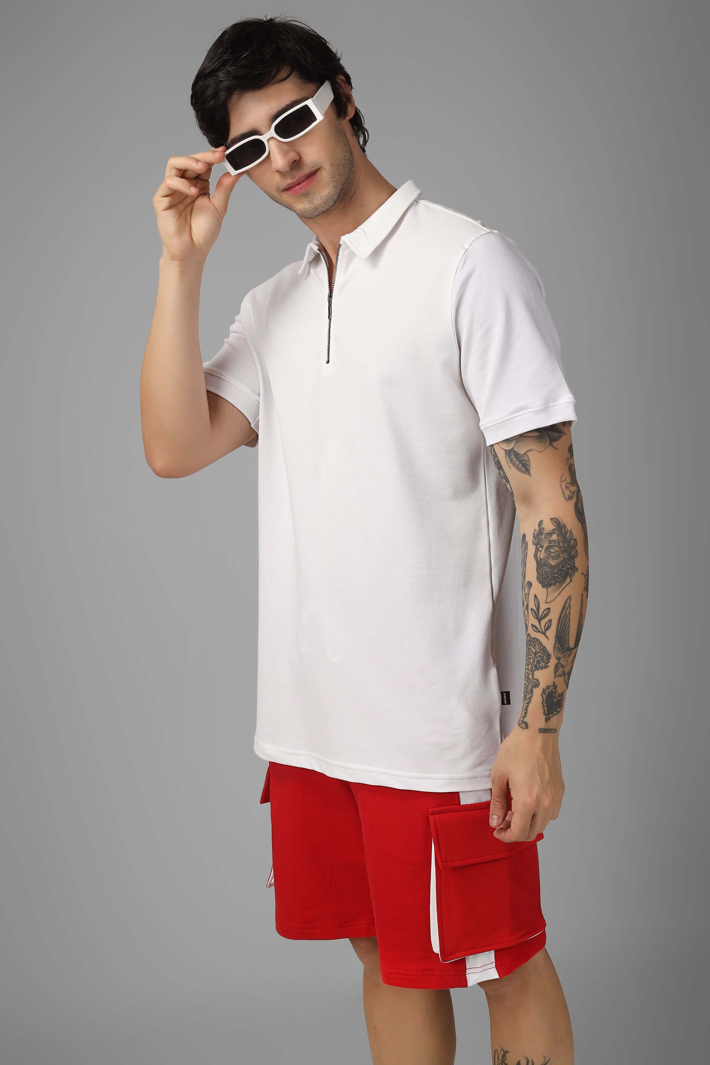 Man wearing a white zipper polo neck t-shirt with sunglasses, paired with red shorts, against a grey background.