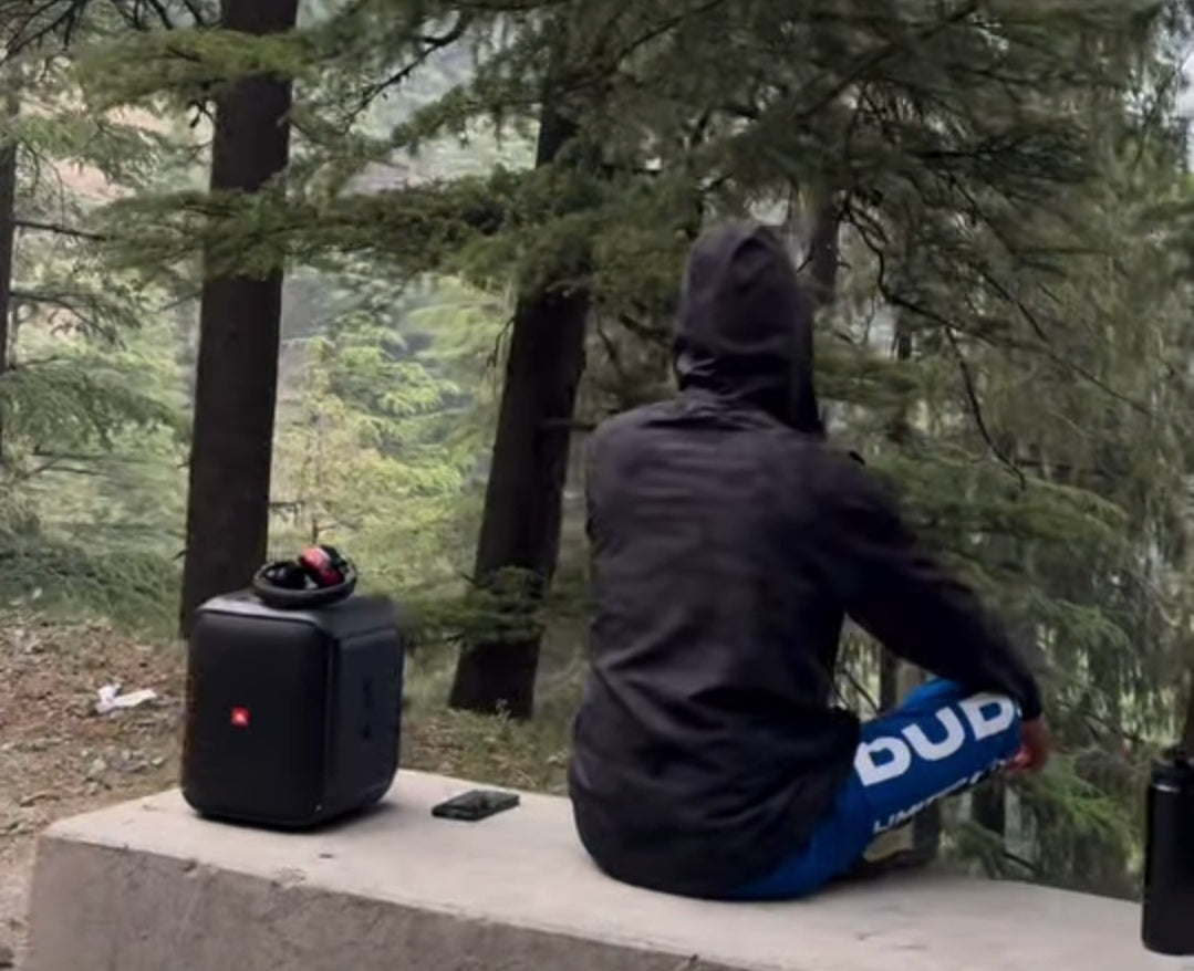 Kapil Sharma wearing blue Duds joggers sitting on a bench in a forest with a speaker nearby.