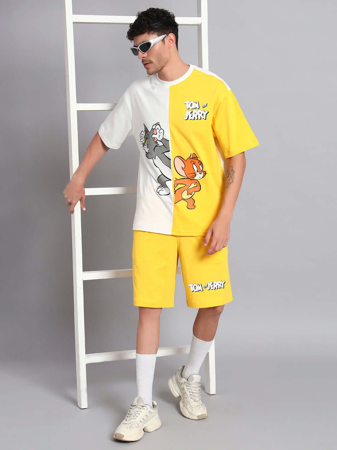 Man wearing a two-tone off-white and yellow Tom & Jerry T-shirt, paired with matching shorts and sunglasses, standing by a ladder.