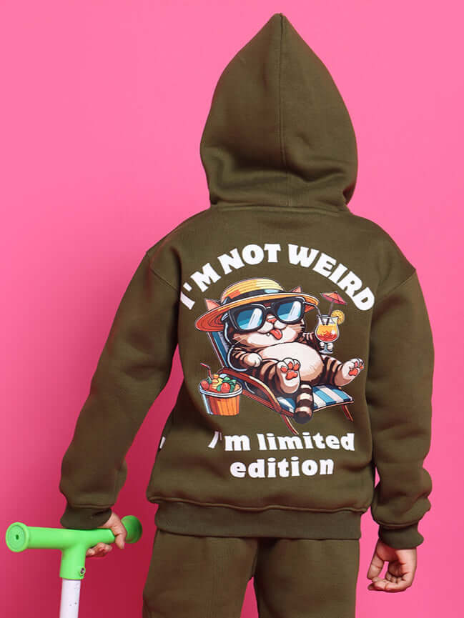 Child in olive green hoodie with cartoon cat design, holding a scooter, against pink background. Text: "I'm not weird, I'm limited edition."