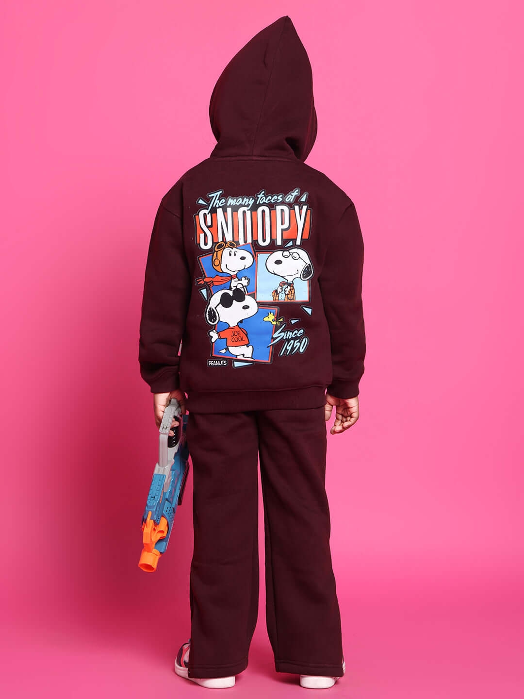 Child wearing Snoopy-themed co-ord set in wine color, featuring a graphic on the back, standing against a pink background, holding a toy.