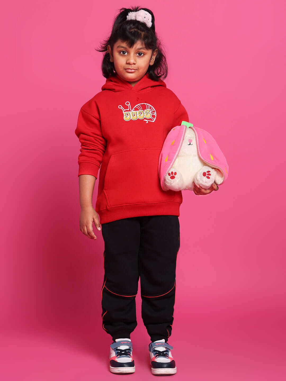 Child in red hoodie and Snail relaxed fit black joggers, holding a plush toy, standing against a pink background.