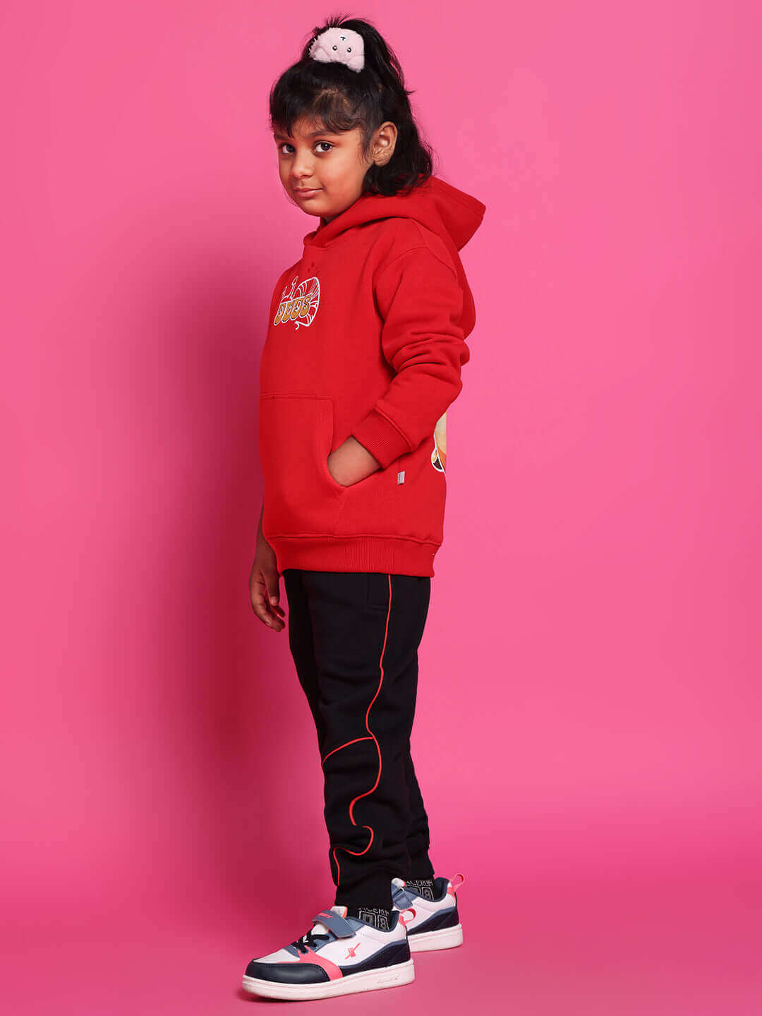 Child wearing Snail Relaxed Fit Jogger in black, suitable for boys and girls, with red hoodie, standing on pink background.