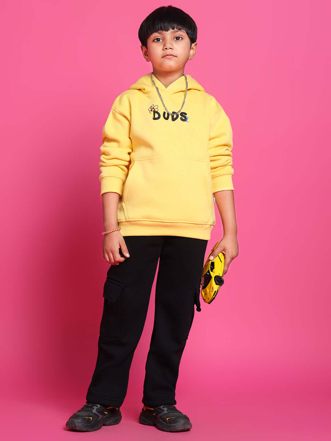 Child wearing COOL DUDE yellow co-ord set with hoodie, black pants, holding a small yellow bag, standing against a pink background.