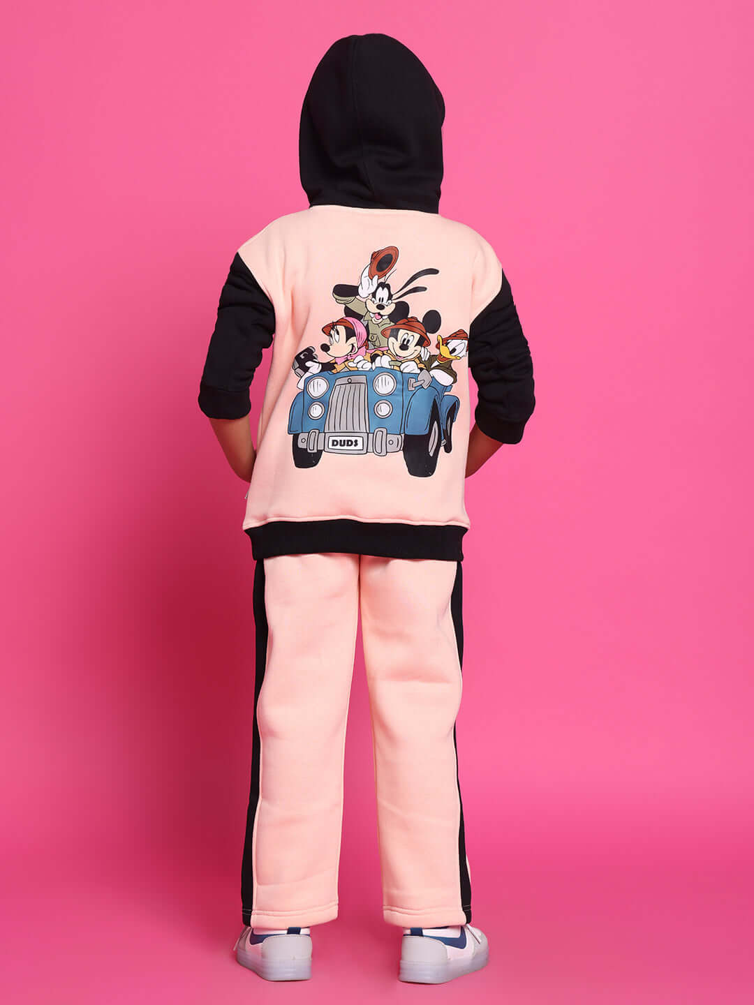 Child wearing Club House co-ord set in black and beige with cartoon print on the back, posing against a pink background.