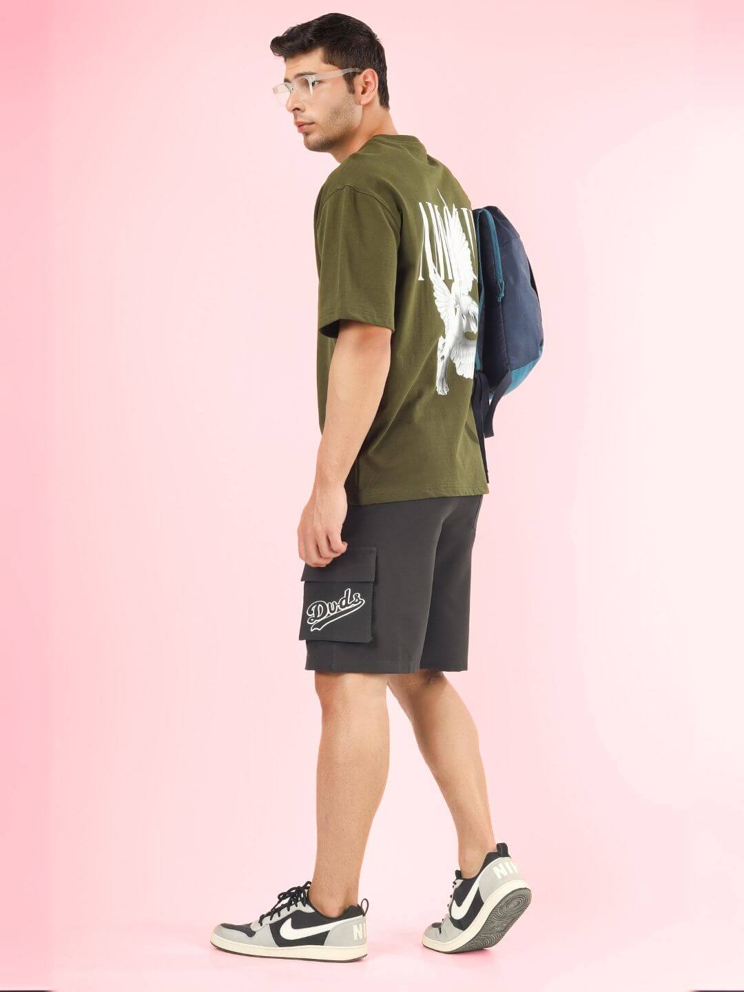Model wearing AMOUR OVER-SIZED T-SHIRT OLIVE GREEN made of premium heavy gauge fabric with black shorts against a pink background.