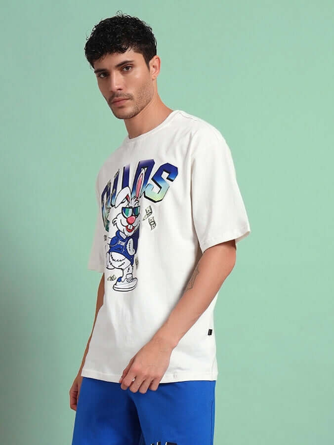 Model wearing Bunny Bugs oversized t-shirt in off-white, styled with blue shorts, against a green background. Trendy summer fashion look.
