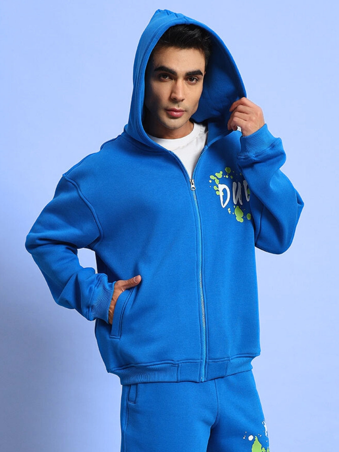 Model wearing blue No Drama cotton fleece hoodie with hood up, featuring artistic splashes and zipper detail.