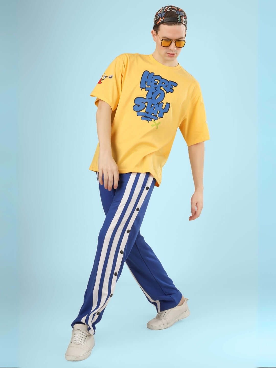 Model wearing HERE TO STAY OVER-SIZED T-SHIRT YELLOW with bold graphic, pairing with striped pants and sunglasses for street style look.