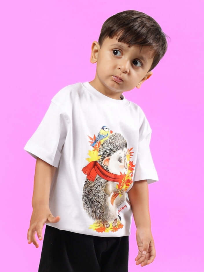 A young child wearing a white hedgehog t-shirt by DUDS, designed for boys and girls, against a bright pink background.
