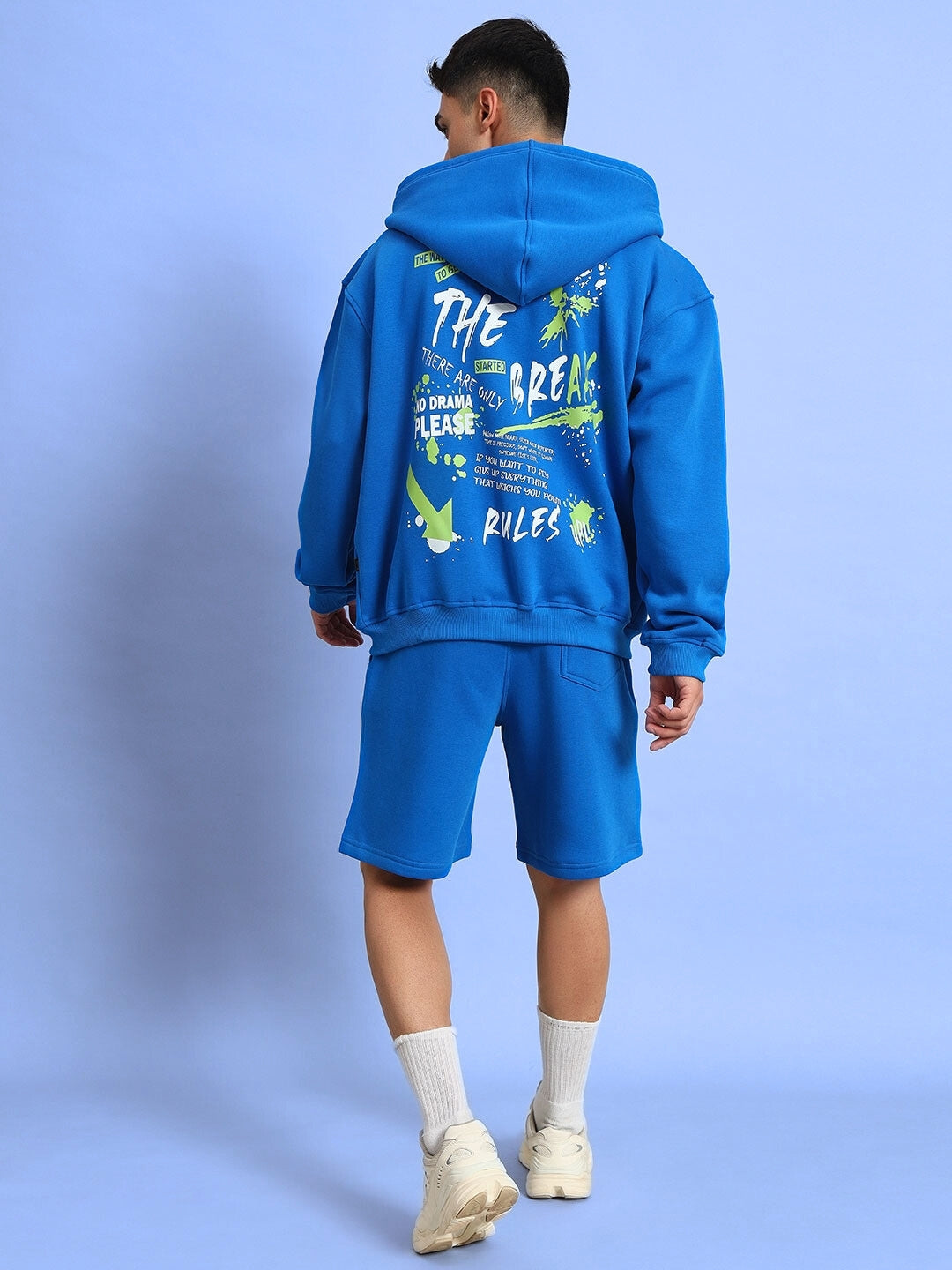 Man wearing blue No Drama Fleece Co-Ord set with graphic back design, featuring hoodie and shorts, on a light blue background.