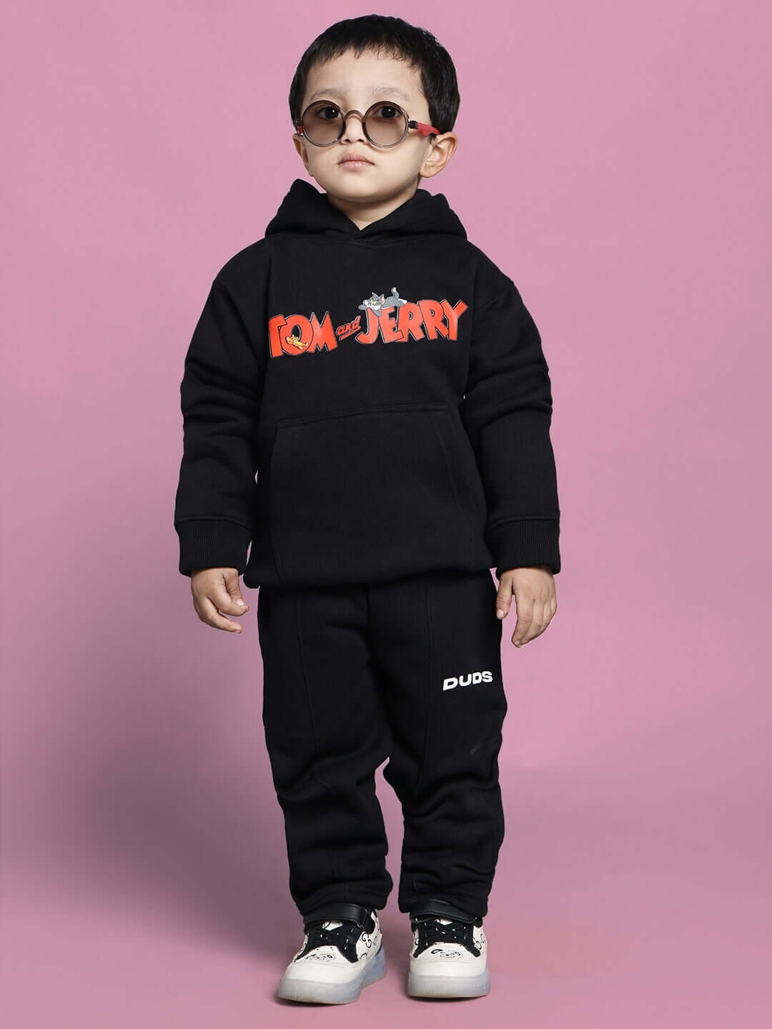 Child wearing black Tom & Jerry co-ord set with hooded sweatshirt and pants, styled with sunglasses, on pink background.