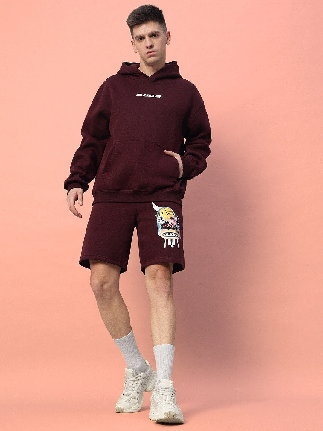 Model wearing JUMBLED FLEECE CO-ORD WINE with graphic print, featuring a hoodie and shorts, against a peach background.