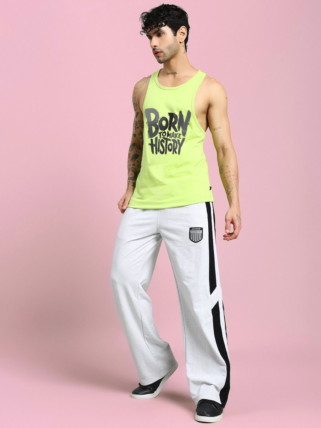 Model wearing BORN TO MAKE HISTORY GYM VEST T-SHIRT NEON GREEN, paired with white pants, standing against a pink background.