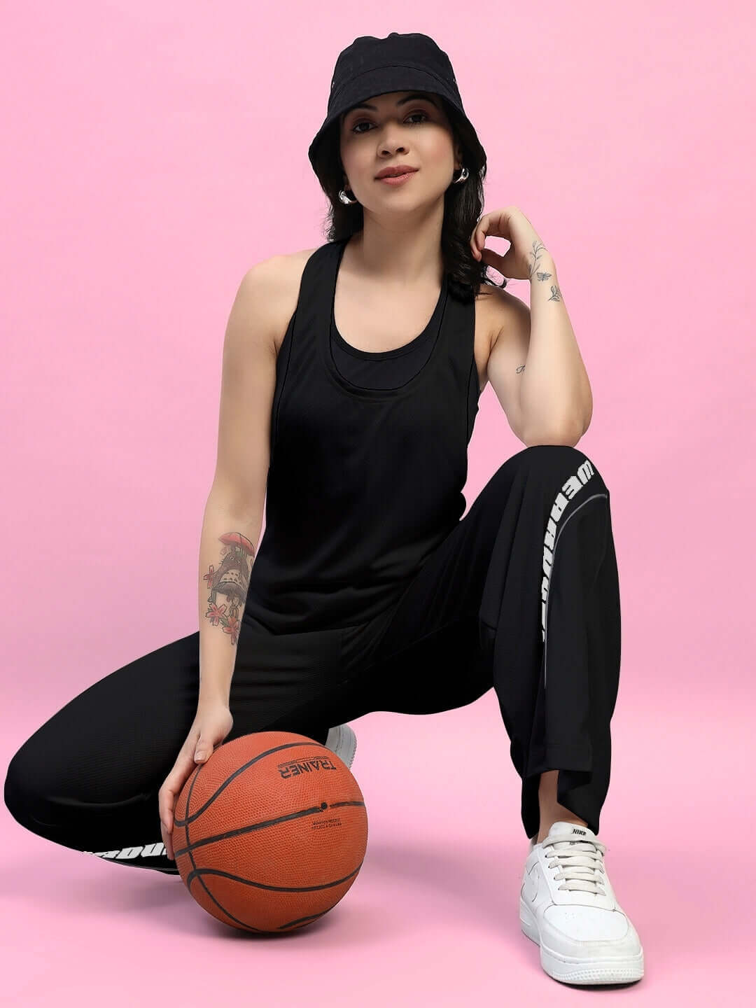 Woman in black joggers and tank top with a basketball, posing against a pink background.