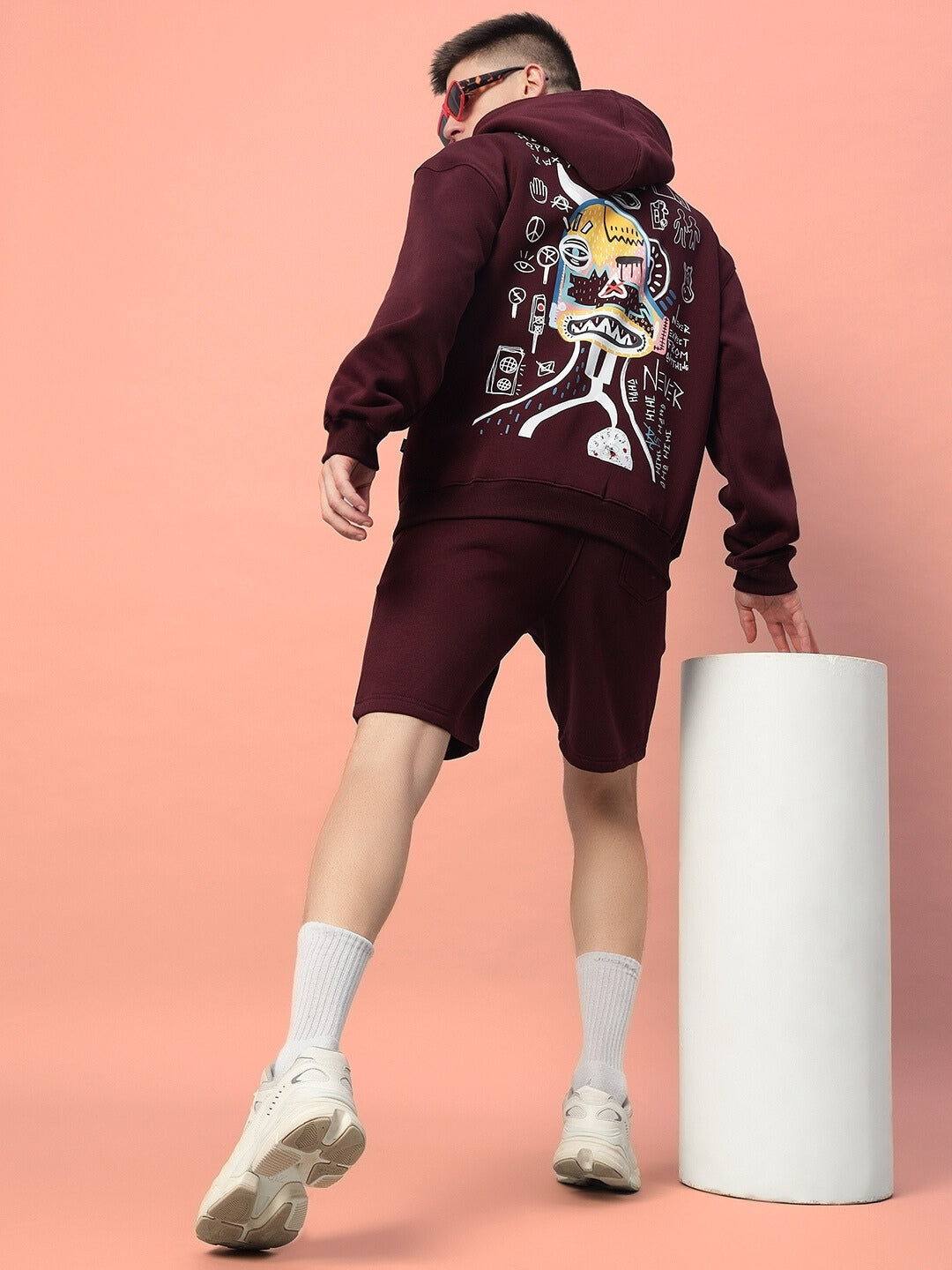Man wearing wine-colored jumbled fleece co-ord with artistic graphic on back, standing against a peach background.