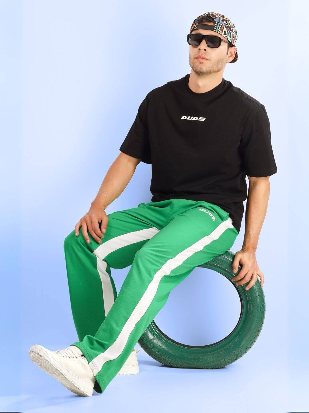 ENJOY THE WIND OVER-SIZED T-SHIRT BLACK worn by a person sitting on a tire, paired with green pants and sunglasses.