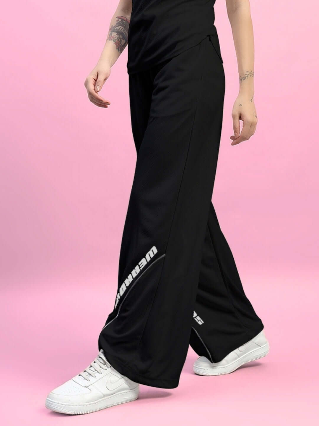 Model wearing Women's Cubby Relaxed Fit Baggy Joggers in black, showcasing wide leg design with white sneakers against pink background.