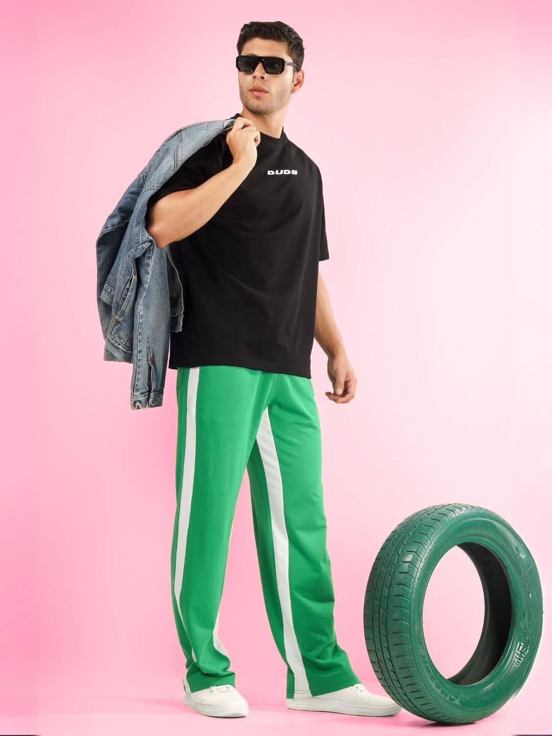 Model wearing ENJOY THE WIND OVER-SIZED T-SHIRT BLACK with green pants, holding denim jacket, standing near tire with pink background.