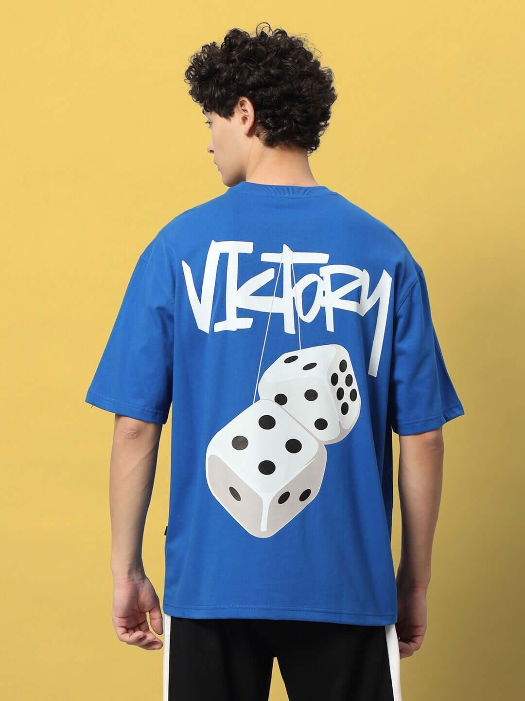 Man wearing VICTORY OVER-SIZED T-SHIRT ROYAL BLUE with dice graphic on the back, facing away against yellow background
