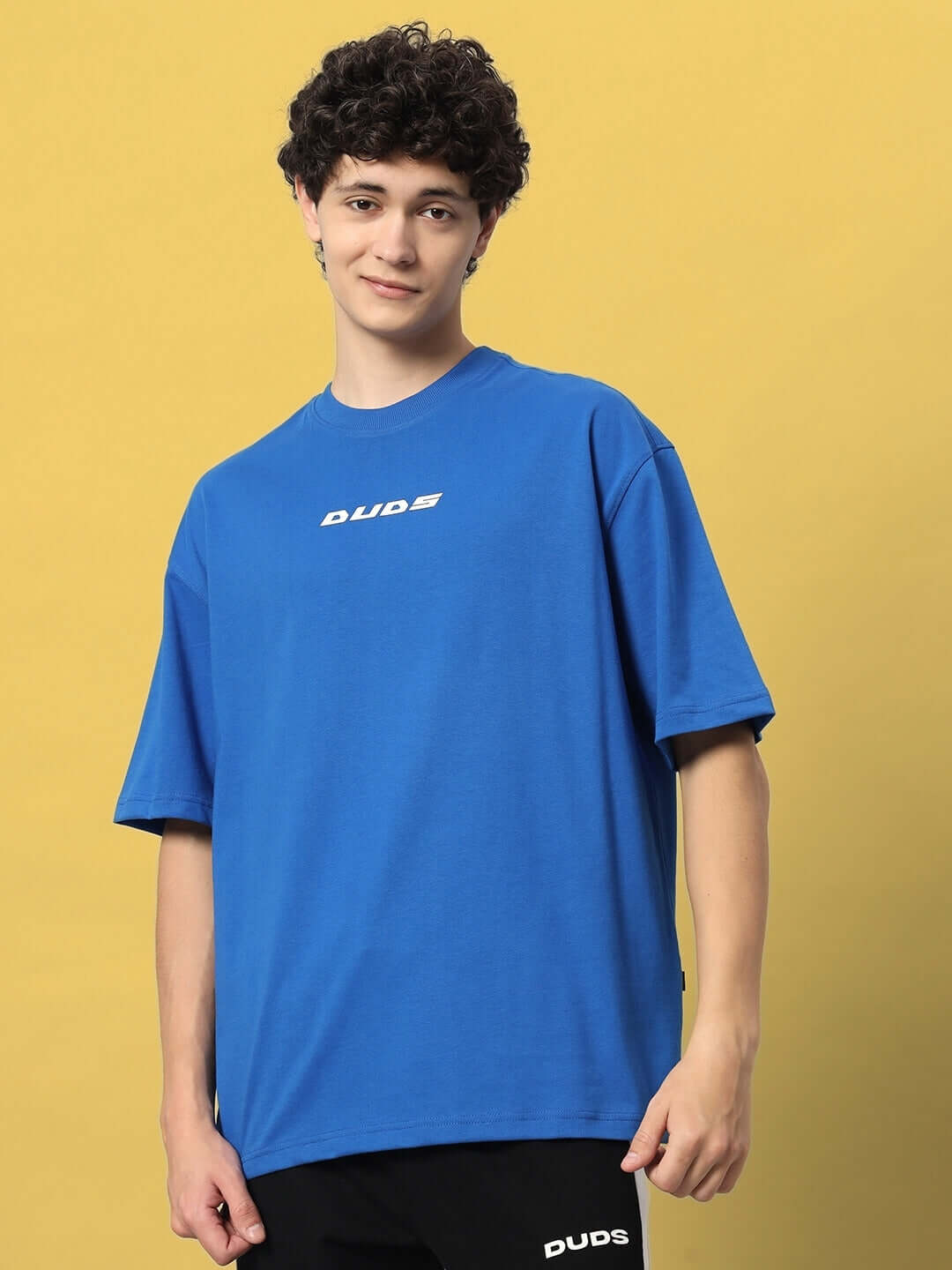 VICTORY OVER-SIZED T-SHIRT ROYAL BLUE worn by a male model against a yellow background, featuring DUDS branding.