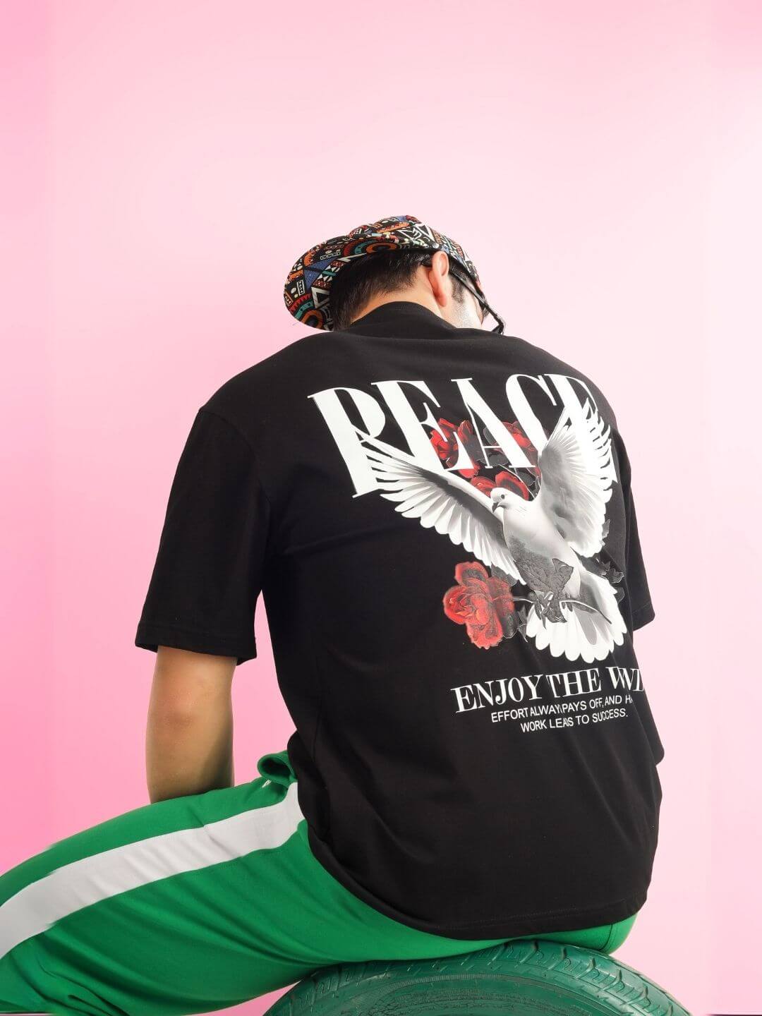 Man wearing ENJOY THE WIND OVER-SIZED T-SHIRT BLACK with dove graphic, against pink background