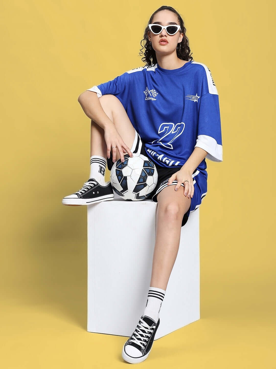 Woman in royal blue sports co-ord set sitting on cube with soccer ball, wearing sunglasses and black sneakers, against yellow background.