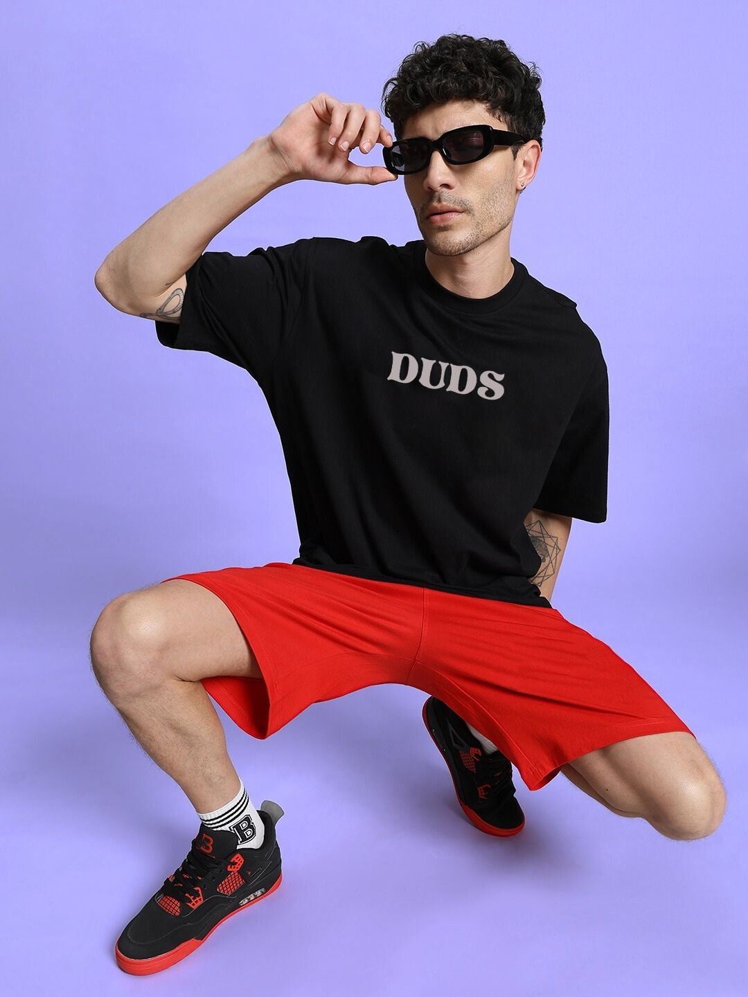Man posing in a black t-shirt and red shorts with sunglasses on a purple background.